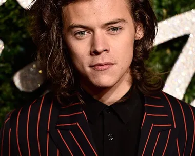 Harry Styles in a red and black striped suit, standing in front of a green foliage background with glittering decorations.