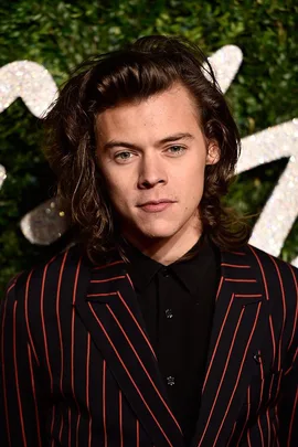 Harry Styles in a red and black striped suit, standing in front of a green foliage background with glittering decorations.