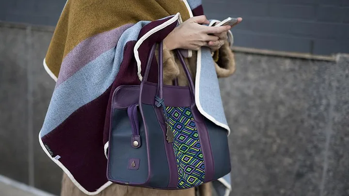 Person using a smartphone is carrying a stylish, multi-colored handbag and wearing a striped blanket wrap.