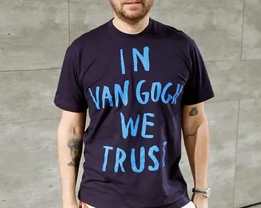 Man wearing a "In Van Gogh We Trust" shirt and "SUP" pants standing against a concrete wall background.