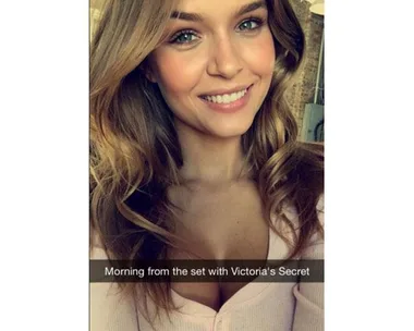 Smiling woman on a Victoria's Secret set, wearing a light pink top, with loose wavy hair and natural makeup.