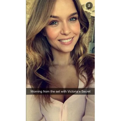 Smiling woman on a Victoria's Secret set, wearing a light pink top, with loose wavy hair and natural makeup.