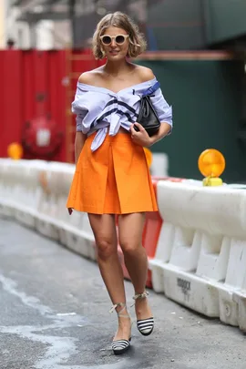 Woman in off-shoulder blue shirt, orange skirt, striped espadrilles, and sunglasses walking on urban street.