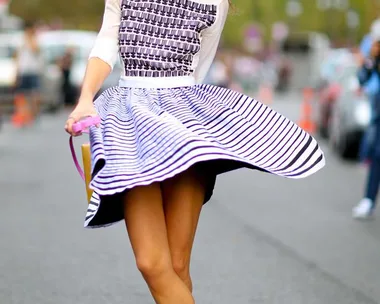 A woman twirls in a patterned dress and white sneakers, smiling and wearing sunglasses on a bustling street.