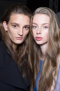 Two models with long brown hair pose closely, showcasing a clean and natural makeup look.