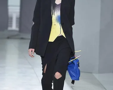 Model walking in Christopher Kane SS16 show wearing black blazer, yellow top, torn pants, and blue shoes, carrying a bag.