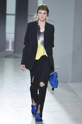 Model walking in Christopher Kane SS16 show wearing black blazer, yellow top, torn pants, and blue shoes, carrying a bag.