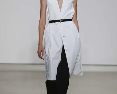 Model in a sleeveless white button-down dress with black pants and belt, walking on the runway.