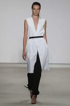 Model in a sleeveless white button-down dress with black pants and belt, walking on the runway.