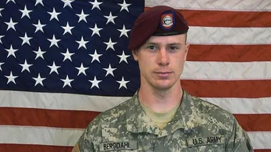 A U.S. Army soldier in uniform stands in front of an American flag, wearing a maroon beret.