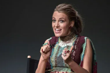 A woman in a colorful dress with a playful expression, seated and gesturing with her hands.