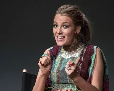 A woman in a colorful dress with a playful expression, seated and gesturing with her hands.