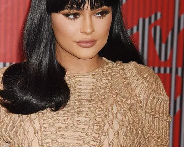 A woman with long black hair and bangs wears a lacy beige top, standing in front of a backdrop with red and gray stripes.