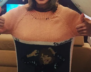 A woman wearing a sweater with an image of herself on it gives a thumbs-up and smiles widely inside an airplane.