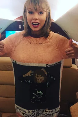 A woman wearing a sweater with an image of herself on it gives a thumbs-up and smiles widely inside an airplane.