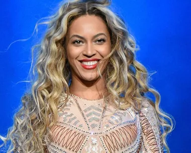 Singer Beyoncé performs at 2015 Made in America festival; she wears a sheer, embellished outfit with wavy, blonde hair and a bright smile.