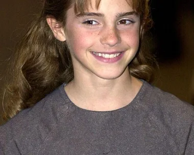 Young girl with long brown hair smiling, wearing a gray dress, pictured in a candid moment.