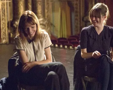 Two women are sitting in a theater, laughing and looking at each other, with empty seats in the background.