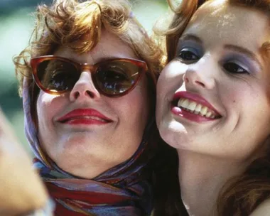 Two women smiling; one with sunglasses and scarf, the other with curly hair and makeup from the movie "Thelma & Louise."