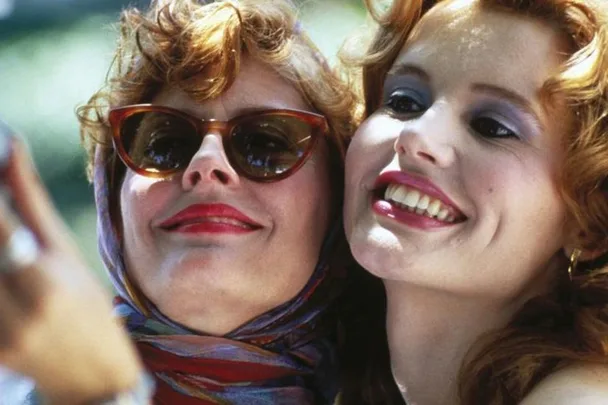 Two women smiling; one with sunglasses and scarf, the other with curly hair and makeup from the movie "Thelma & Louise."