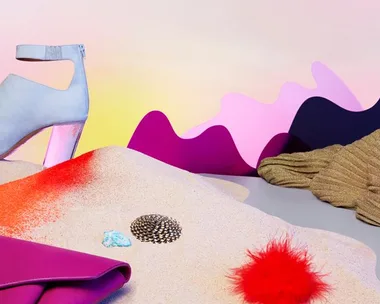 A blue high-heeled shoe, pink handbag, textured patterns, sand, and a red fluffy accessory on a colorful abstract background.