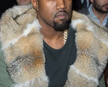 Person looking serious, wearing a fur-collared jacket while seated at a fashion event, with people in the background.