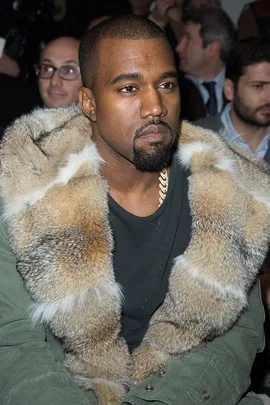 Person looking serious, wearing a fur-collared jacket while seated at a fashion event, with people in the background.