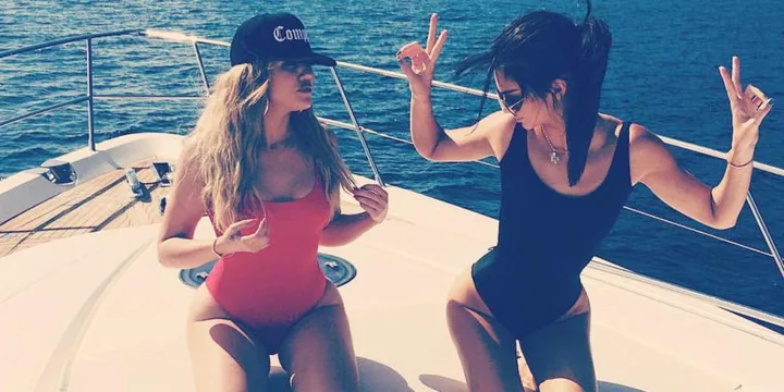 Two women in swimsuits pose playfully on a yacht under the sun, with the ocean in the background.