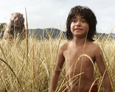 Mowgli, a shirtless young boy with dark hair, stands in tall grass with a tiger blurred in the background in "The Jungle Book" (2016).