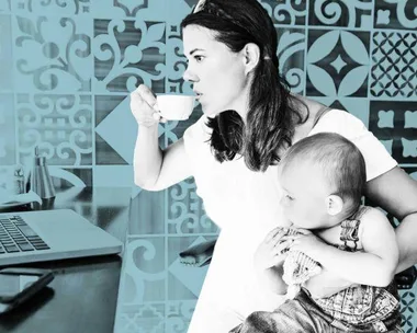 Mother holding baby while working on laptop and having a drink.