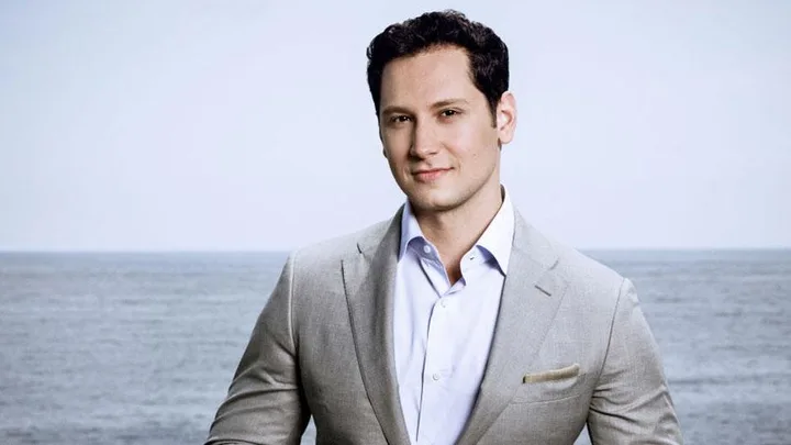 A man in a light gray suit stands in front of the ocean, smiling slightly at the camera.