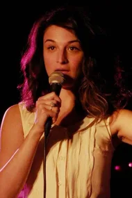 Woman with wavy hair speaking into a microphone under a spotlight.