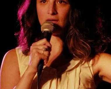 Woman with wavy hair speaking into a microphone under a spotlight.