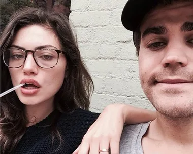 A couple taking a casual selfie against a brick wall, the woman wearing glasses and the man in a cap.