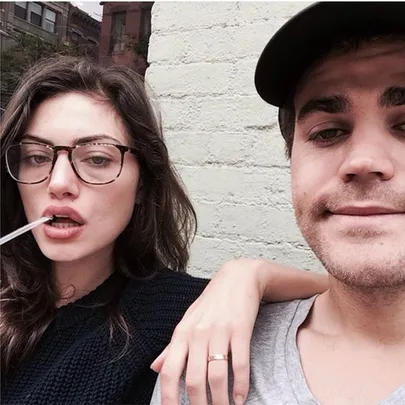 A couple taking a casual selfie against a brick wall, the woman wearing glasses and the man in a cap.