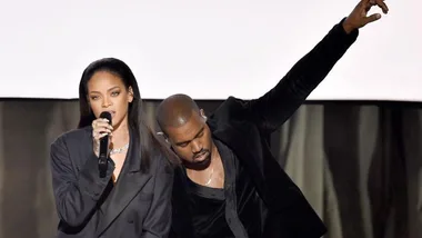 Rihanna singing into a microphone next to Kanye West on stage, with Kanye raising his arm dressed in black outfits.