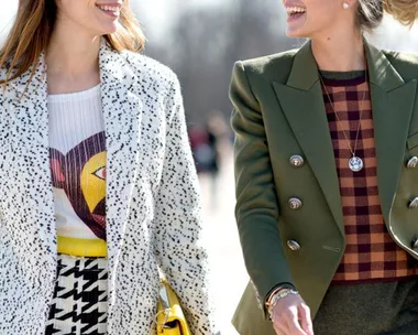 Two women are walking together, smiling and wearing stylish outfits. One in a white speckled coat, the other in a green blazer.