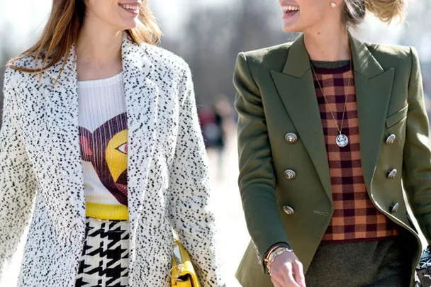 Two women are walking together, smiling and wearing stylish outfits. One in a white speckled coat, the other in a green blazer.