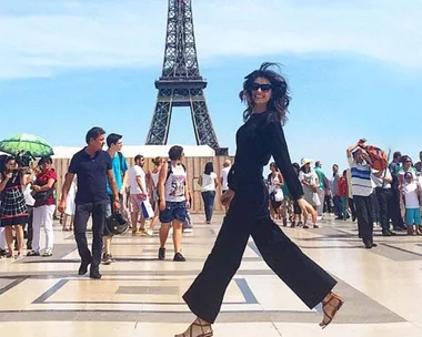 Woman in black jumpsuit smiling, walking near Eiffel Tower with crowd in background.