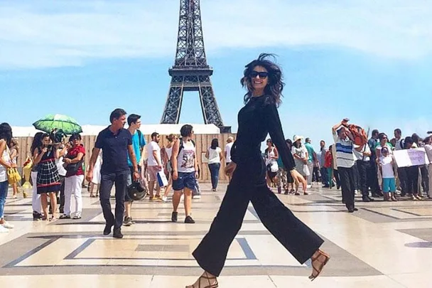 Woman in black jumpsuit smiling, walking near Eiffel Tower with crowd in background.