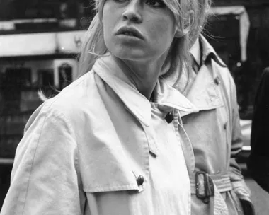 A woman with a blonde beehive hairstyle and trench coat stands outdoors, gazing to the left with a serious expression.