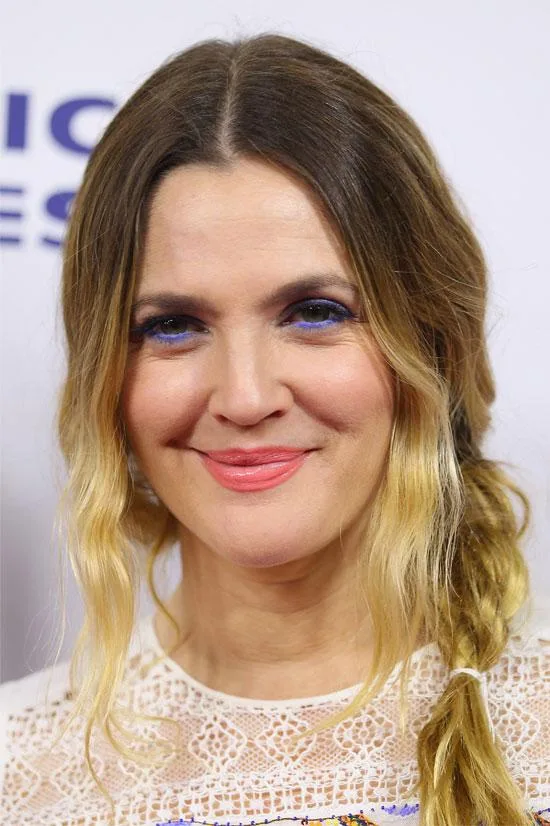 A woman with blue eyeliner, coral lipstick, and a side braid smiles against a light background.