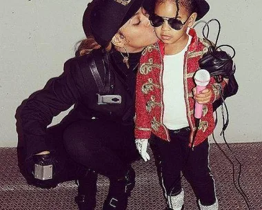 Parent in black costume kisses child dressed as Michael Jackson with red jacket and fedora, holding a microphone.