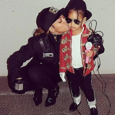 Parent in black costume kisses child dressed as Michael Jackson with red jacket and fedora, holding a microphone.