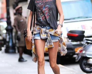 Woman in Iron Maiden shirt, denim shorts, boots; flannel tied at waist, walking on street.