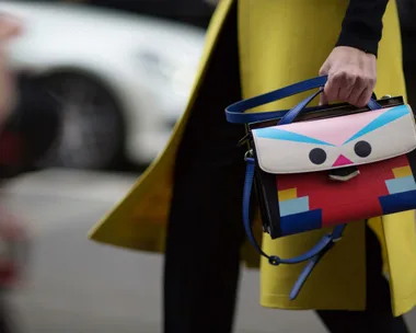 A person holds a colorful handbag with a fun geometric face design, wearing a yellow coat, on a city street background.