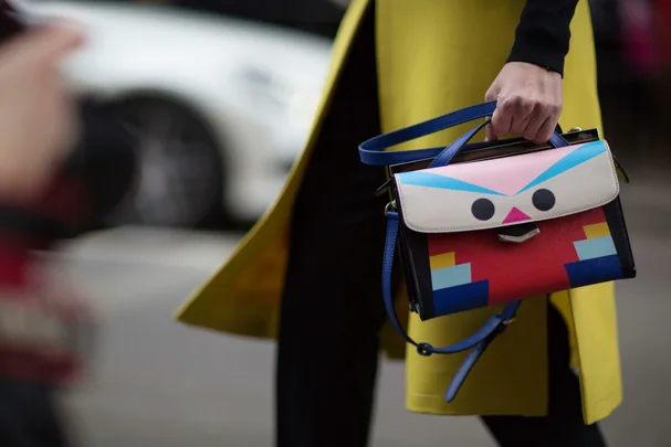 A person holds a colorful handbag with a fun geometric face design, wearing a yellow coat, on a city street background.