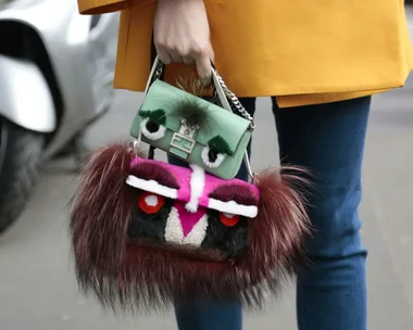 Fashionable woman holding two colorful, furry handbags designed with eye-like details, wearing a yellow coat.