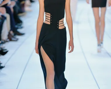 Model walking down runway in a black, high-neck dress with side straps and thigh-high slit.