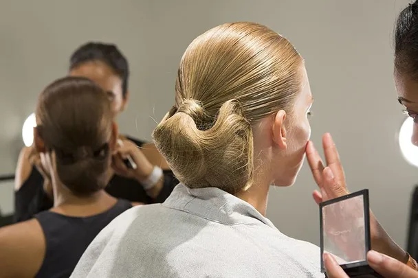 Backstage beauty preparation, woman with sleek bun getting makeup applied by artist under bright lights.